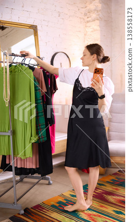 Woman trying outfit in mirror while using smartphone at home. Woman trying outfit in mirror while using smartphone at home. 138558173