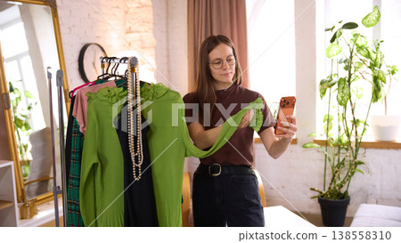 Woman holding green garment and smartphone comparing clothes before purchase Woman holding green garment and smartphone comparing clothes before purchase 138558310