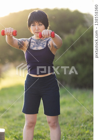 Tattooed young woman exercising with dumbbells outdoors 138558360