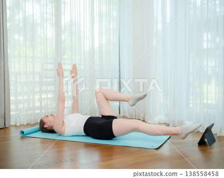 Young woman doing core exercise at home on yoga mat. Concept of fitness, strength training, and online workout. Female performing alternating leg raise while following virtual class. Young woman doing core exercise at home on yoga mat. Concept of fitness, strength training, and online workout. Female performing alternating leg raise while following virtual class. 138558458