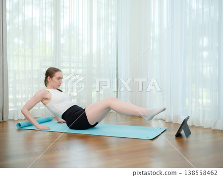 Young woman doing core exercise at home on yoga mat. Concept of fitness, strength training, and online workout. Female performing leg raise while following virtual fitness class. Young woman doing core exercise at home on yoga mat. Concept of fitness, strength training, and online workout. Female performing leg raise while following virtual fitness class. 138558462