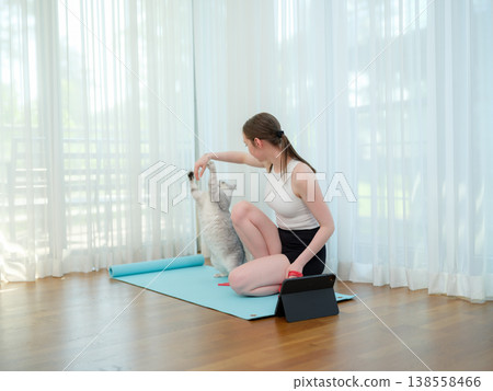 Young woman resting after workout at home with her cat. Concept of fitness, pet companionship, and healthy lifestyle. Female relaxing on yoga mat and playing with domestic animal. 138558466