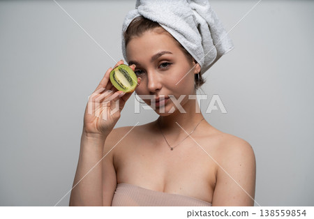 Woman with a towel on her head holding a half kiwi fruit next to her eye, representing natural skincare, beauty, wellness, organic ingredients, and healthy lifestyle Woman with a towel on her head holding a half kiwi fruit next to her eye, representing natural skincare, beauty, wellness, organic ingredients, and healthy lifestyle 138559854