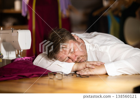 Late in evening, tired man tailor dozed off next to sewing machine. Late in evening, tired man tailor dozed off next to sewing machine. 138560121