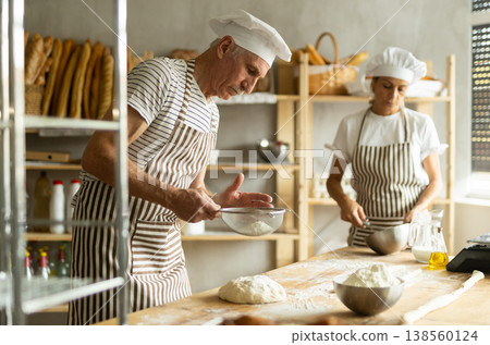 Two bakers prepare dough for baguettes - whisking eggs and sprinkling flour over the dough through sieve Two bakers prepare dough for baguettes - whisking eggs and sprinkling flour over the dough through sieve 138560124