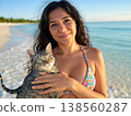 A bikini-clad woman, holding her beloved cat and beaming with a sunny smile, enjoys a tropical vacation against the backdrop of a crystal-clear blue ocean. 138560287