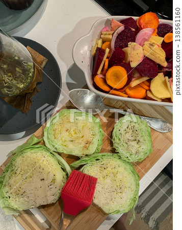 A collection of vegetables is placed in a baking dish for cooking in the oven. Chopped cabbage sits on a cutting board with a brush and cooking tools near the vegetables A collection of vegetables is placed in a baking dish for cooking in the oven. Chopped cabbage sits on a cutting board with a brush and cooking tools near the vegetables 138561160