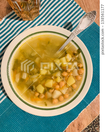 Bowl holds a warm potato and chickpea soup. The soup has chunks of potato and chickpeas in broth. A spoon rests in the bowl. Nearby is a glass filled with a drink Bowl holds a warm potato and chickpea soup. The soup has chunks of potato and chickpeas in broth. A spoon rests in the bowl. Nearby is a glass filled with a drink 138561177