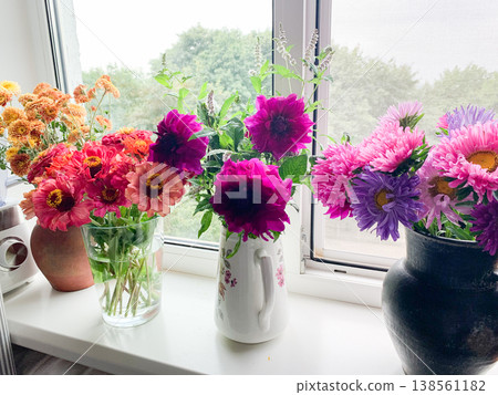 A vase with seasonal flowers sits on a windowsill in an apartment. Sunlight shines through the window, highlighting the colorful blooms and greenery 138561182