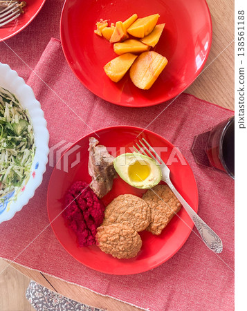 Breakfast is served on red plates with turkey breast, beetroot hummus, fresh salad, and carrot buns. A peach is included for dessert, creating a balanced meal for the day Breakfast is served on red plates with turkey breast, beetroot hummus, fresh salad, and carrot buns. A peach is included for dessert, creating a balanced meal for the day 138561188