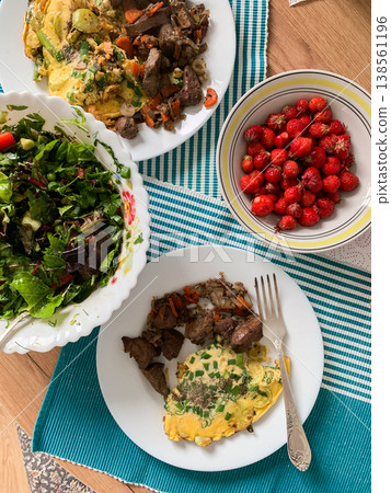 A meal features beef liver with a zucchini omelette on a white plate, fresh salad in a bowl, and strawberries for dessert on a table with a blue and white tablecloth A meal features beef liver with a zucchini omelette on a white plate, fresh salad in a bowl, and strawberries for dessert on a table with a blue and white tablecloth 138561196