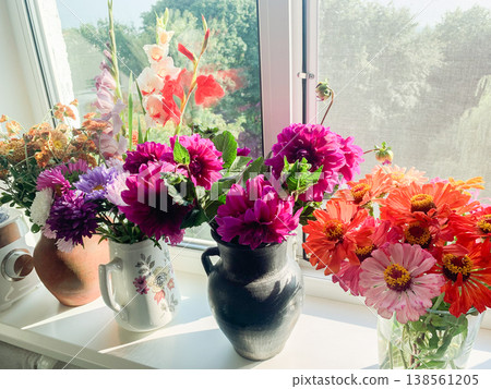 A vase with seasonal flowers sits on a windowsill in an apartment. Sunlight shines through the window, highlighting the colorful blooms and greenery 138561205