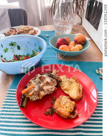 A healthy breakfast includes an omelette with carrots, chicken breast, and green beans, served on a red plate. A salad and fruit sit on the table, with natural light coming in 138561208