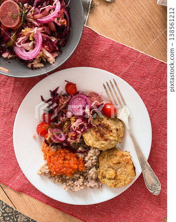 A healthy breakfast plate includes green buckwheat, chicken cutlets, zucchini caviar, and a side salad with radish and tomatoes. Fresh ingredients are showcased on a simple table setting 138561212