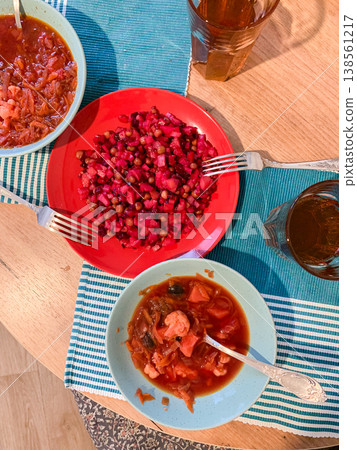 A bowl of Ukrainian borscht sits next to a plate of colorful salad and another bowl of food on a wooden table. Glasses filled with a beverage are placed around the dishes A bowl of Ukrainian borscht sits next to a plate of colorful salad and another bowl of food on a wooden table. Glasses filled with a beverage are placed around the dishes 138561217