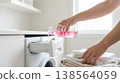 Person pours pink liquid detergent into laundry machine drawer while folded towels are stacked on countertop. Bright room with natural light coming from window. 138564059