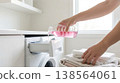 Person pours laundry detergent from bottle into washing machine drawer. Bright laundry room with clean towels folded on countertop. Concept of cleaning, home care, laundry service. 138564061