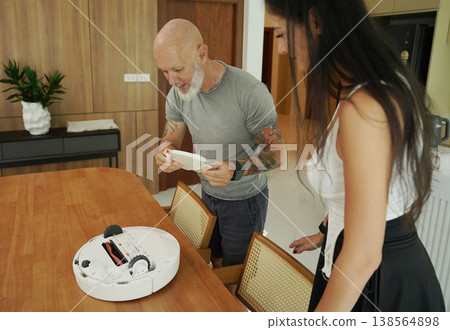 A man and a woman clean a robotic vacuum cleaner in a bright living room. A man and a woman clean a robotic vacuum cleaner in a bright living room. 138564898