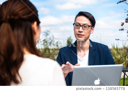 Businessmen and women having a meeting on a terrace, gathered around laptops. Businessmen and women having a meeting on a terrace, gathered around laptops. 138565778