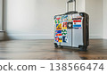 silver suitcase covered in travel stickers from around the world, placed on an empty wooden floor with white walls in the background 138566474