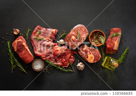 Different cuts of beef steak are arranged on a black surface. Fresh herbs, spices, and garlic accompany the meat, ready for cooking or grilling at a kitchen or market 138566685