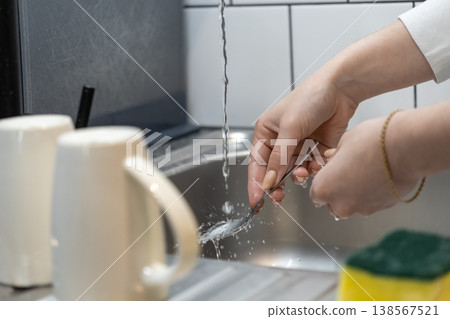 A person's hands meticulously wash a shiny metal spoon under running water in a modern kitchen sink, highlighting daily chores and cleanliness A person's hands meticulously wash a shiny metal spoon under running water in a modern kitchen sink, highlighting daily chores and cleanliness 138567521