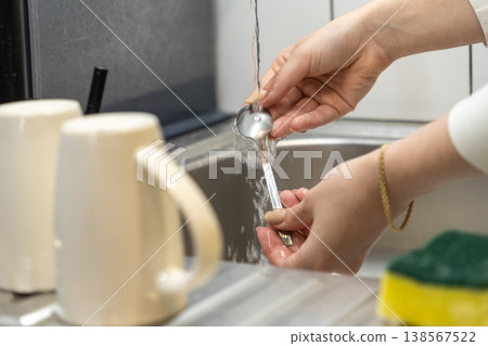 Female hands washing a metal spoon under running water in a modern kitchen sink, depicting essential daily household chores and maintaining cleanliness 138567522