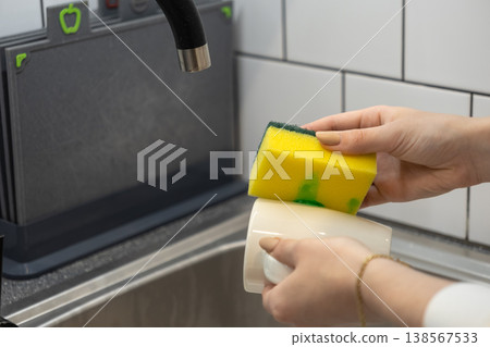 Person's hands vigorously washing a white porcelain mug with a bright yellow scrubbing sponge and dish soap, demonstrating daily home cleaning and essential kitchen hygiene Person's hands vigorously washing a white porcelain mug with a bright yellow scrubbing sponge and dish soap, demonstrating daily home cleaning and essential kitchen hygiene 138567533