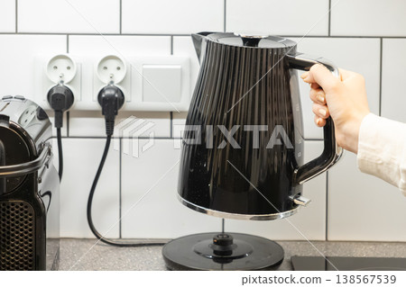 Person's hand carefully lifting a sleek black electric kettle, featuring 1.7 L capacity and modern ribbed design, from its base on a kitchen counter Person's hand carefully lifting a sleek black electric kettle, featuring 1.7 L capacity and modern ribbed design, from its base on a kitchen counter 138567539