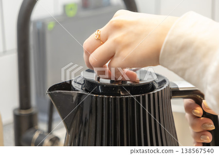 Woman's hand opening the lid of a modern black electric kettle, filling it with water for a hot drink, highlighting a routine for domestic comfort Woman's hand opening the lid of a modern black electric kettle, filling it with water for a hot drink, highlighting a routine for domestic comfort 138567540