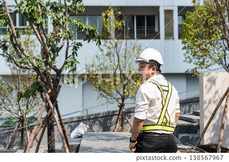 A male engineer in a safety vest surveys an outdoor construction site surrounded by trees. The image combines urban progress with environmental awareness. A male engineer in a safety vest surveys an outdoor construction site surrounded by trees. The image combines urban progress with environmental awareness. 138567967