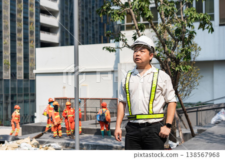 A civil engineer stands confidently overseeing construction workers in the background. The image reflects leadership, awareness, and commitment to project success. A civil engineer stands confidently overseeing construction workers in the background. The image reflects leadership, awareness, and commitment to project success. 138567968