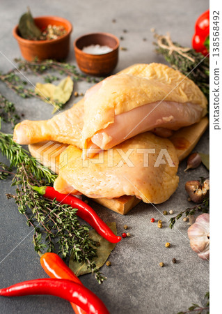 Raw chicken legs rest on a wooden cutting board with fresh herbs and spices placed nearby. The scene takes place in a kitchen with natural light coming in 138568492