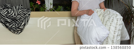 A 53-year-old woman is changing her bed linen in her bedroom. She carefully removes old linen and adds fresh, clean sheets to the bed while standing in a well-lit room, banner A 53-year-old woman is changing her bed linen in her bedroom. She carefully removes old linen and adds fresh, clean sheets to the bed while standing in a well-lit room, banner 138568850