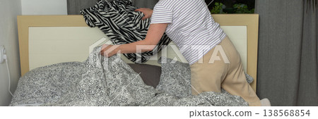 A woman of 53 years old is changing bed linen in her bedroom. She removes old linen and replaces it with new bedding while standing by the bed in daylight, banner 138568854