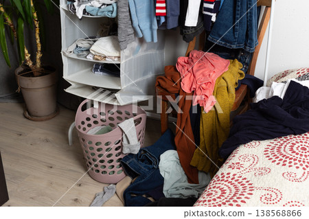 Clothes are scattered on the floor and inside a basket in a room. A chair holds additional clothes. The scene shows items ready for washing after daily use and activities Clothes are scattered on the floor and inside a basket in a room. A chair holds additional clothes. The scene shows items ready for washing after daily use and activities 138568866