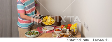 A 53-year-old woman prepares dinner at a round wooden table in her kitchen. She pours food from a colander onto a plate while arranging utensils and other items, banner A 53-year-old woman prepares dinner at a round wooden table in her kitchen. She pours food from a colander onto a plate while arranging utensils and other items, banner 138568910