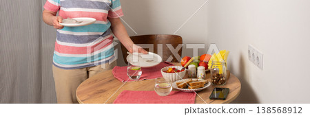 A 53-year-old woman sets the table for dinner. She carries a stack of plates and a glass of water. Fresh fruits and vegetables are arranged on the table, ready for the meal, banner A 53-year-old woman sets the table for dinner. She carries a stack of plates and a glass of water. Fresh fruits and vegetables are arranged on the table, ready for the meal, banner 138568912