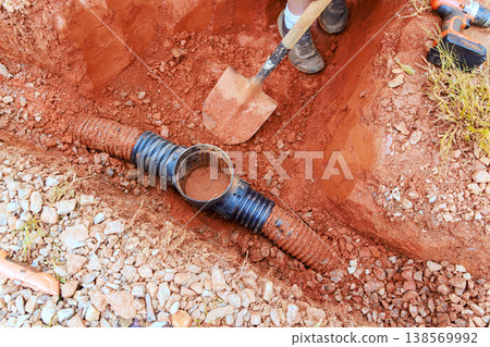 Worker digs trench for drainage pipe installation in red dirt, section of black pipe, tools nearby. Worker digs trench for drainage pipe installation in red dirt, section of black pipe, tools nearby. 138569992