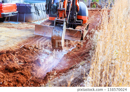 Worker operates excavator digger on dirt road in an outdoor digging trench Worker operates excavator digger on dirt road in an outdoor digging trench 138570024