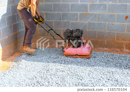 Worker uses plate compactor on gravel in building under construction area has cinder block walls. Worker uses plate compactor on gravel in building under construction area has cinder block walls. 138570104