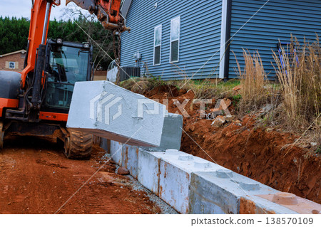 Construction workers use excavator to lift place concrete blocks for retaining walls beside house. 138570109