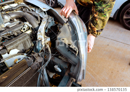 Worker is taking off headlight section of car in garage tools, car parts are scattered around. Worker is taking off headlight section of car in garage tools, car parts are scattered around. 138570135