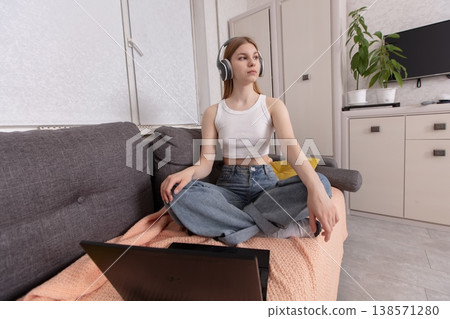 Young woman in casual attire engaging in online activities while sitting cross-legged on a couch in a cozy living room Young woman in casual attire engaging in online activities while sitting cross-legged on a couch in a cozy living room 138571280
