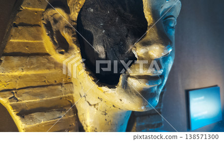 Ancient egyptian pharaoh mask or mummy head exhibit in a museum, featuring a broken face detail with visible inner layers, golden textures, and dramatic lighting. 138571300