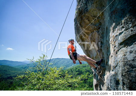 Aerial view of male rock climber ascending rugged limestone cliff with harness and rope for safety. Sportsman climbing on vertical large boulder at Dobvush Rocks in Carpathian mountains, Ukraine. Aerial view of male rock climber ascending rugged limestone cliff with harness and rope for safety. Sportsman climbing on vertical large boulder at Dobvush Rocks in Carpathian mountains, Ukraine. 138571443