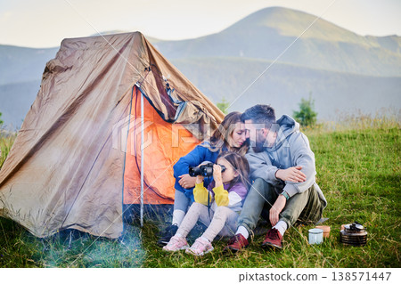 Family of three sits together in front of orange tent in grassy mountain campsite. Child uses binoculars while parents share tender moment. Peaceful and intimate outdoor experience. 138571447