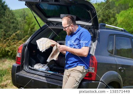 Man tourist leans on open trunk of SUV, deeply focused on map. Traveler wears blue shirt and beige pants, standing in lush, green area, carefully planning or navigating his route. Man tourist leans on open trunk of SUV, deeply focused on map. Traveler wears blue shirt and beige pants, standing in lush, green area, carefully planning or navigating his route. 138571449
