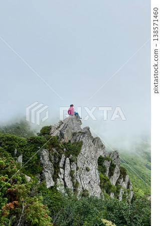 Woman hiker in pink jacket and blue pants sits atop jagged rock outcrop, surrounded by mist and lush greenery. Foggy atmosphere adds sense of mystery and tranquility to dramatic mountain landscape. 138571460