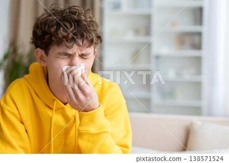 Young teen guy in yellow hoodie blowing nose into tissue on sofa Young teen guy in yellow hoodie blowing nose into tissue on sofa 138571524